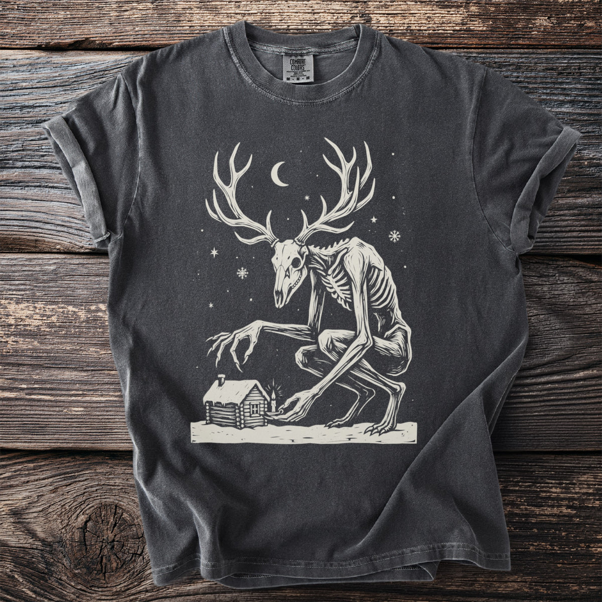 Pepper gray t-shirt with a cream folk-cut print: a gaunt antlered wendigo with a deer skull face stalks toward a lone cabin in the snow.