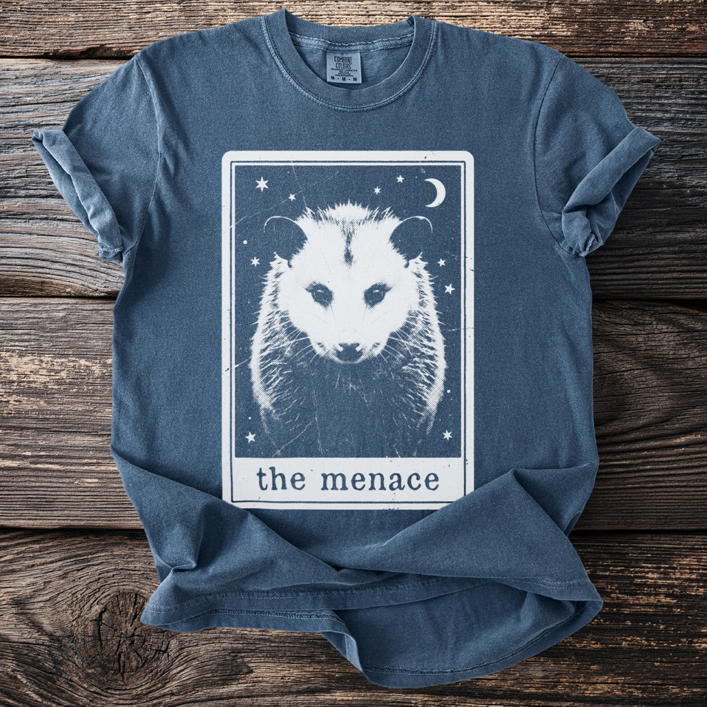 Blue t-shirt with a white graphic of a possum face in a tarot card titled "the menace."