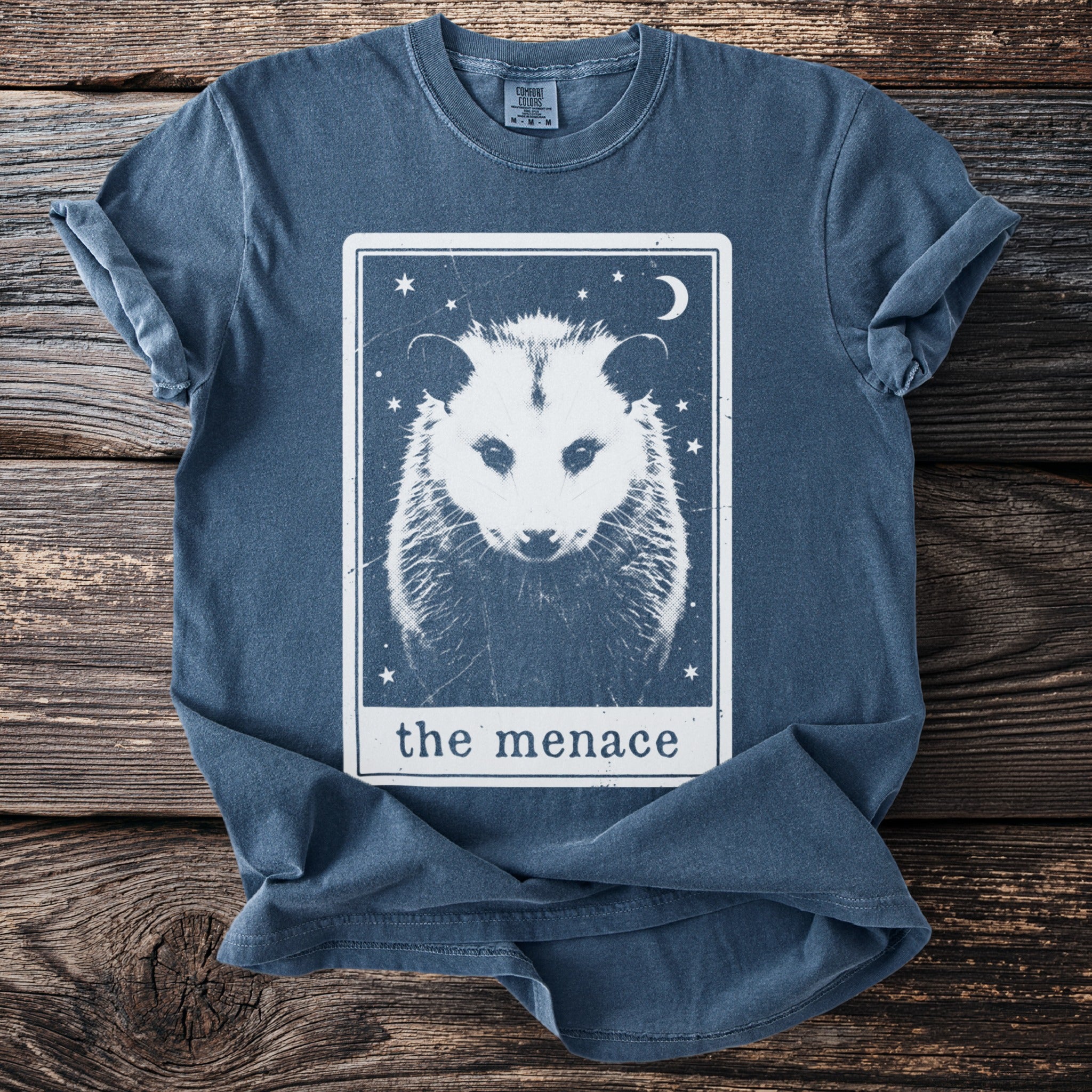 Blue t-shirt with a white graphic of a possum face in a tarot card titled "the menace."