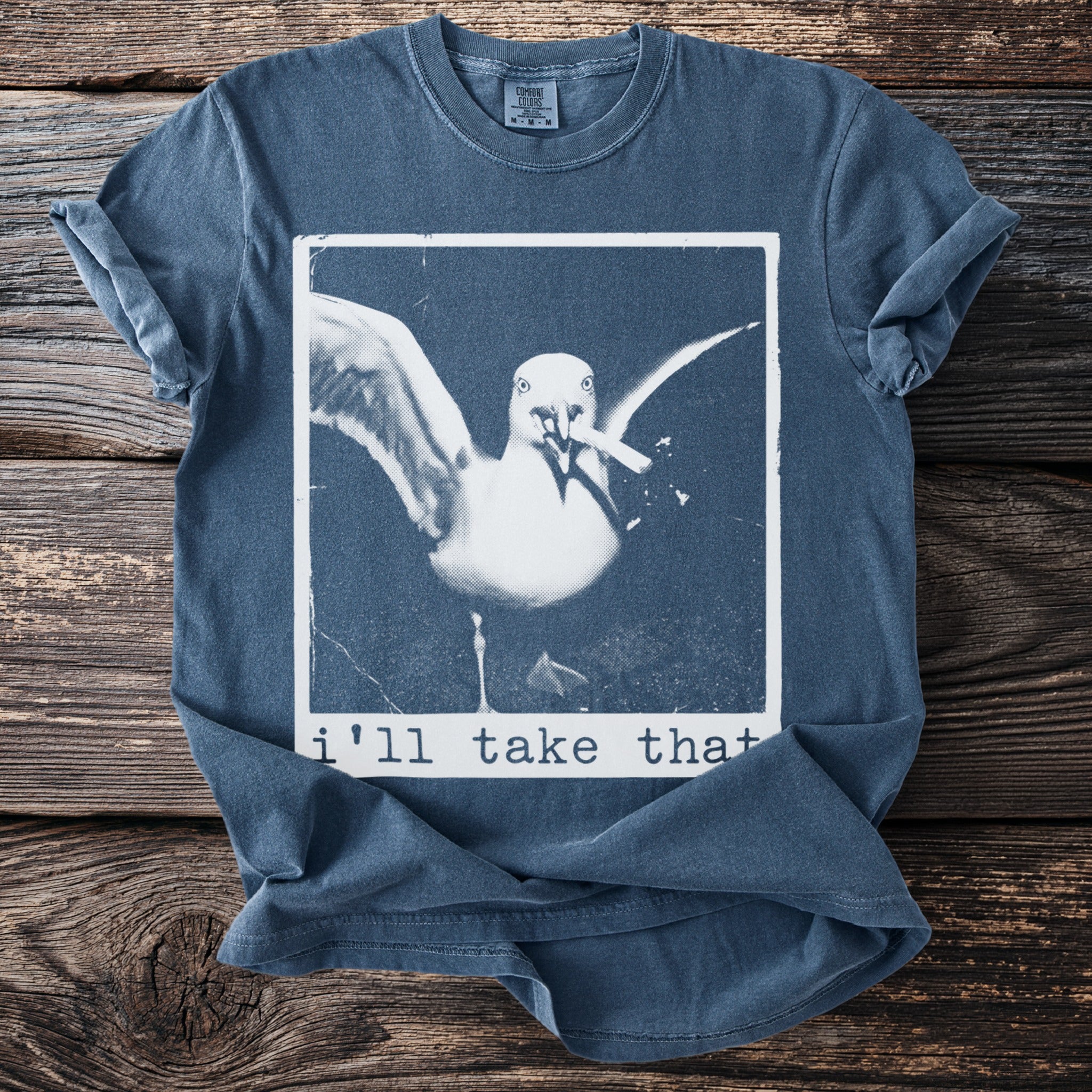 Blue t-shirt with a distressed print of a seagull holding a cigarette that says "i'll take that."