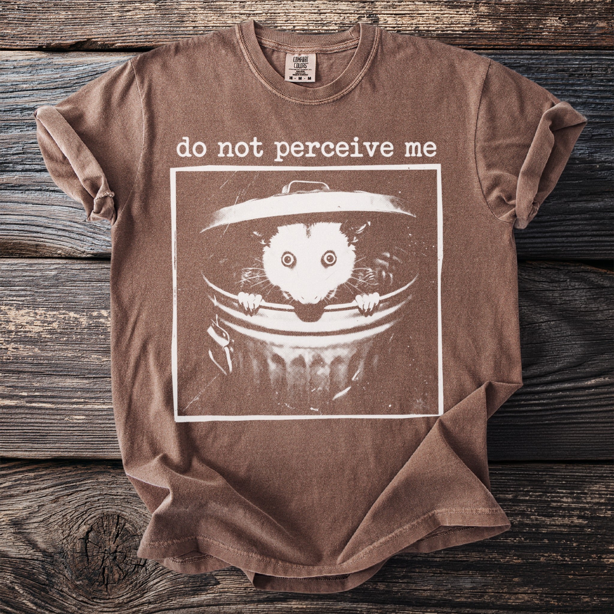 Brown t-shirt with graphic of an opossum in a trash can and text "do not perceive me".