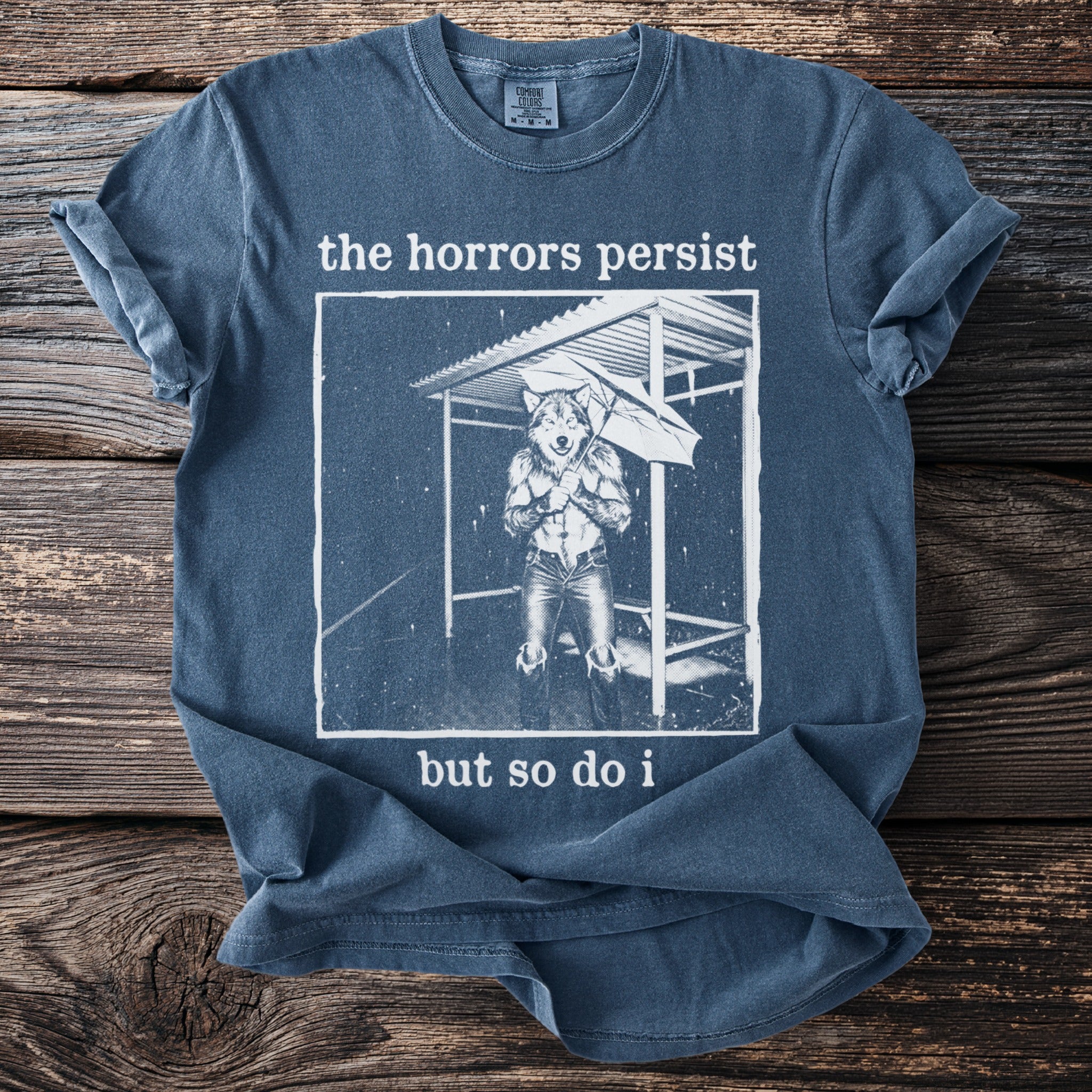 Blue t-shirt with graphic of anthropomorphic wolf holding umbrella, text "the horrors persist but so do i".