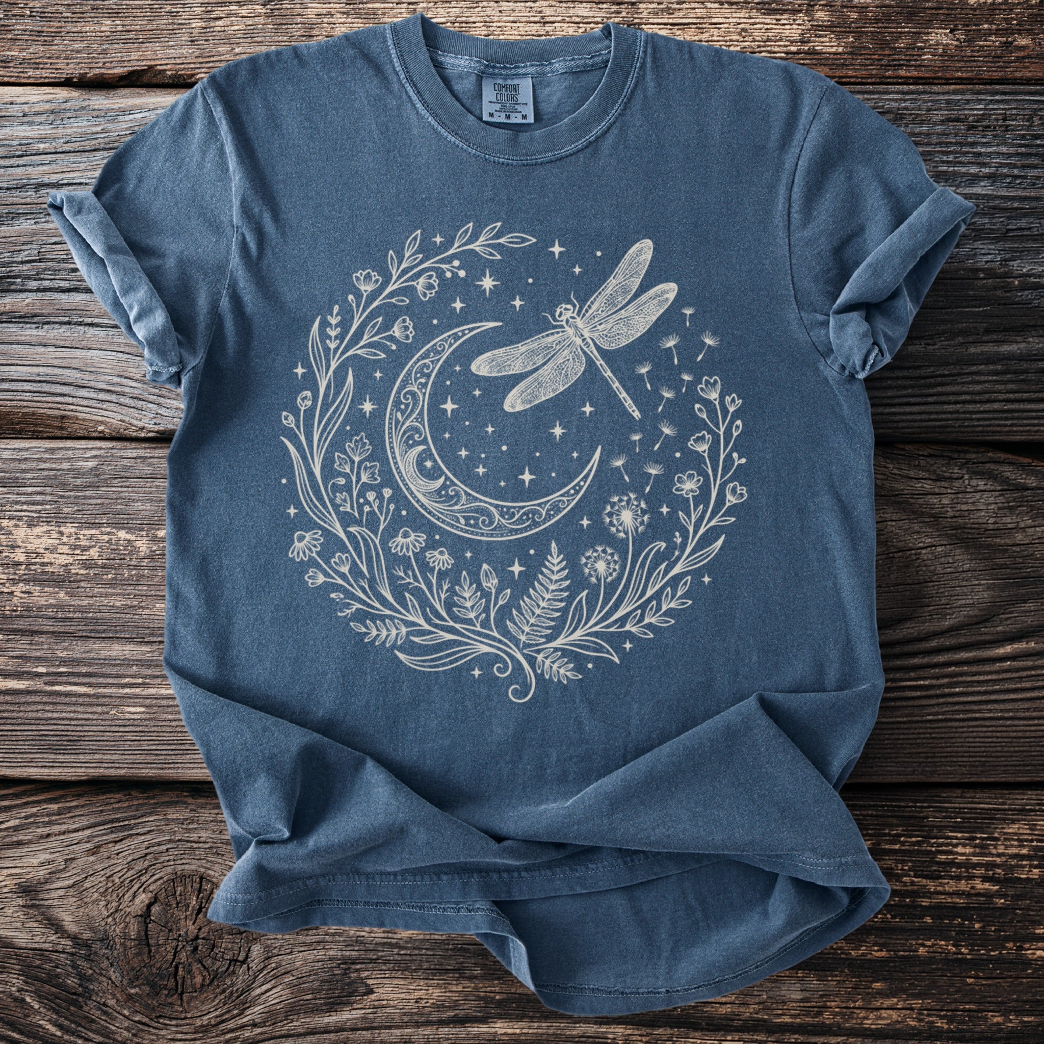 Blue t-shirt with a white line drawing of a crescent moon, dragonfly, stars, and plants.