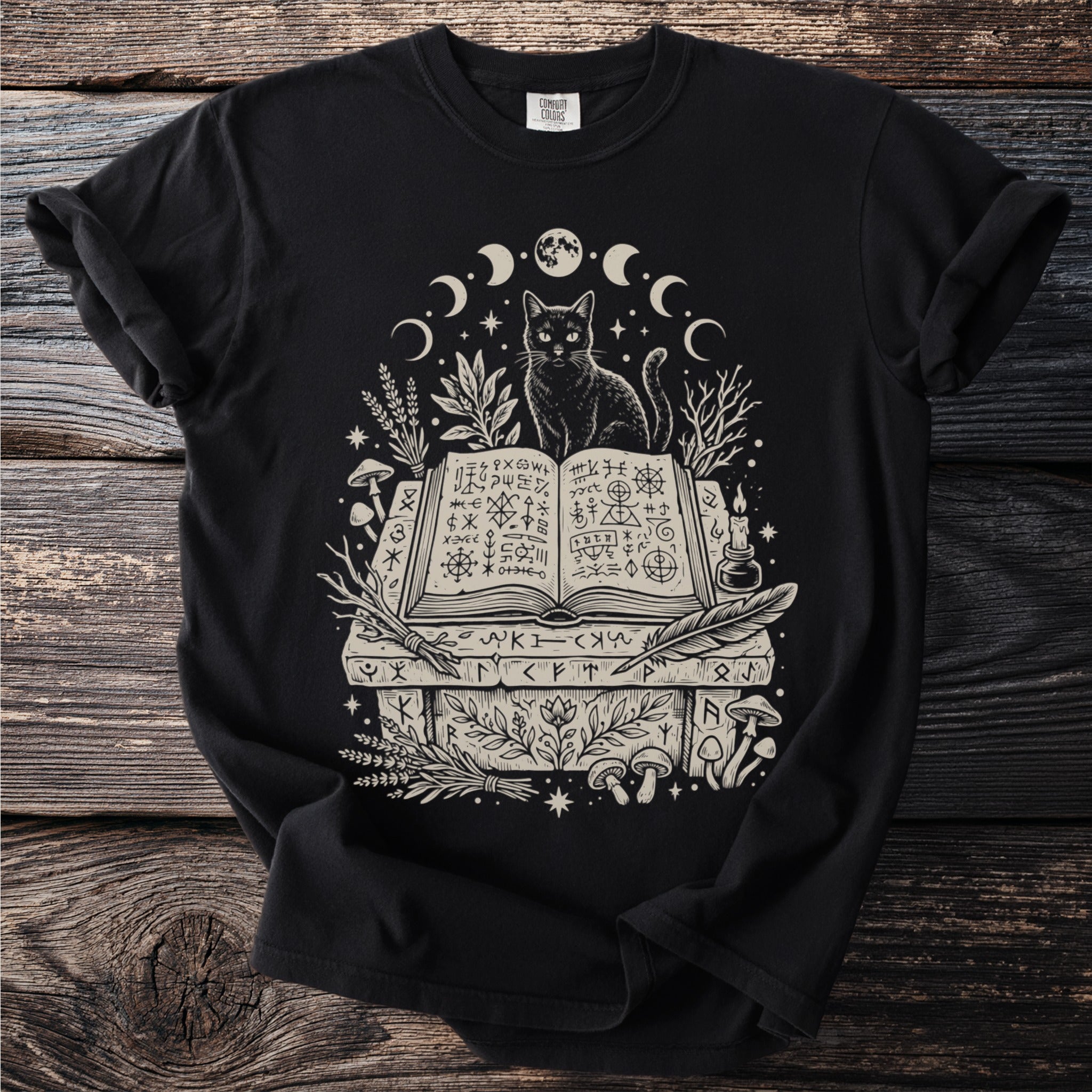 Black t-shirt featuring a black cat sitting on an open book with mystical symbols and celestial elements.