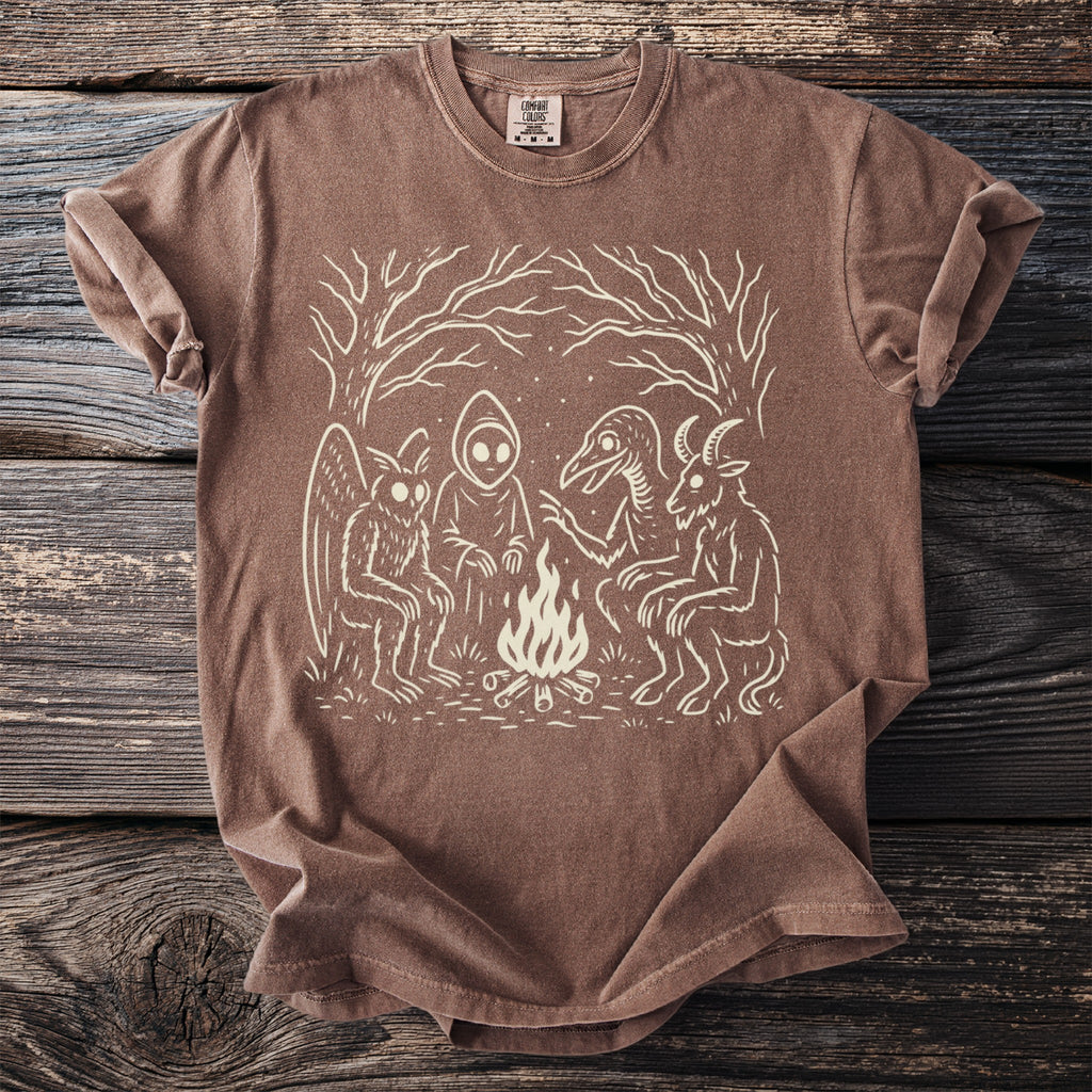 Brown t-shirt with a white graphic of cryptids around a campfire.