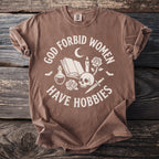 Brown t-shirt with "GOD FORBID WOMEN HAVE HOBBIES" text and occult imagery.