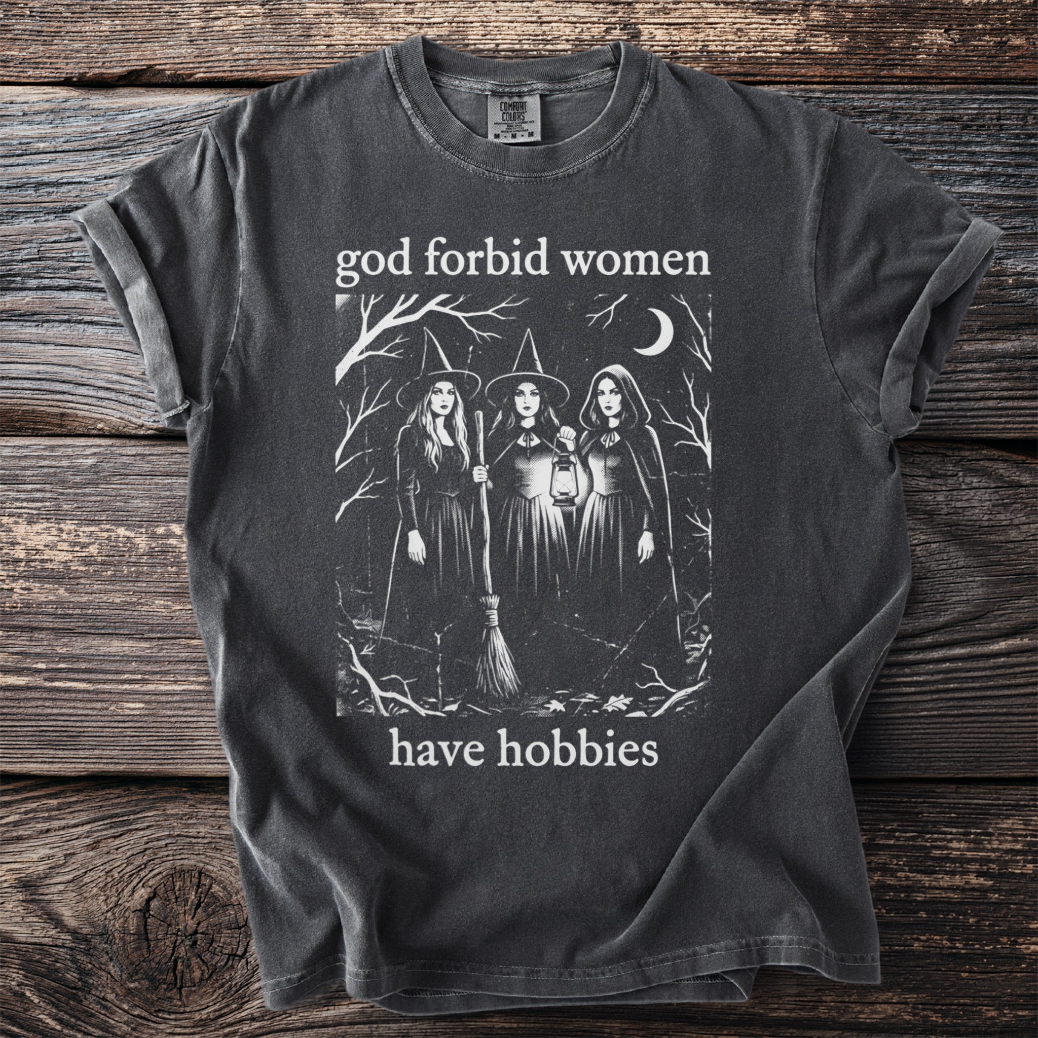 Dark wash t-shirt with "god forbid women have hobbies" and three witches illustration.