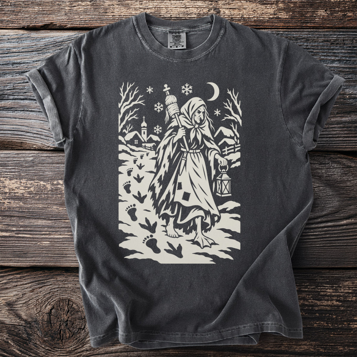 Pepper gray t-shirt with a cream folk-cut print: Perchta walks through a snowy village with a lantern and a spindle, goose footprints behind her.