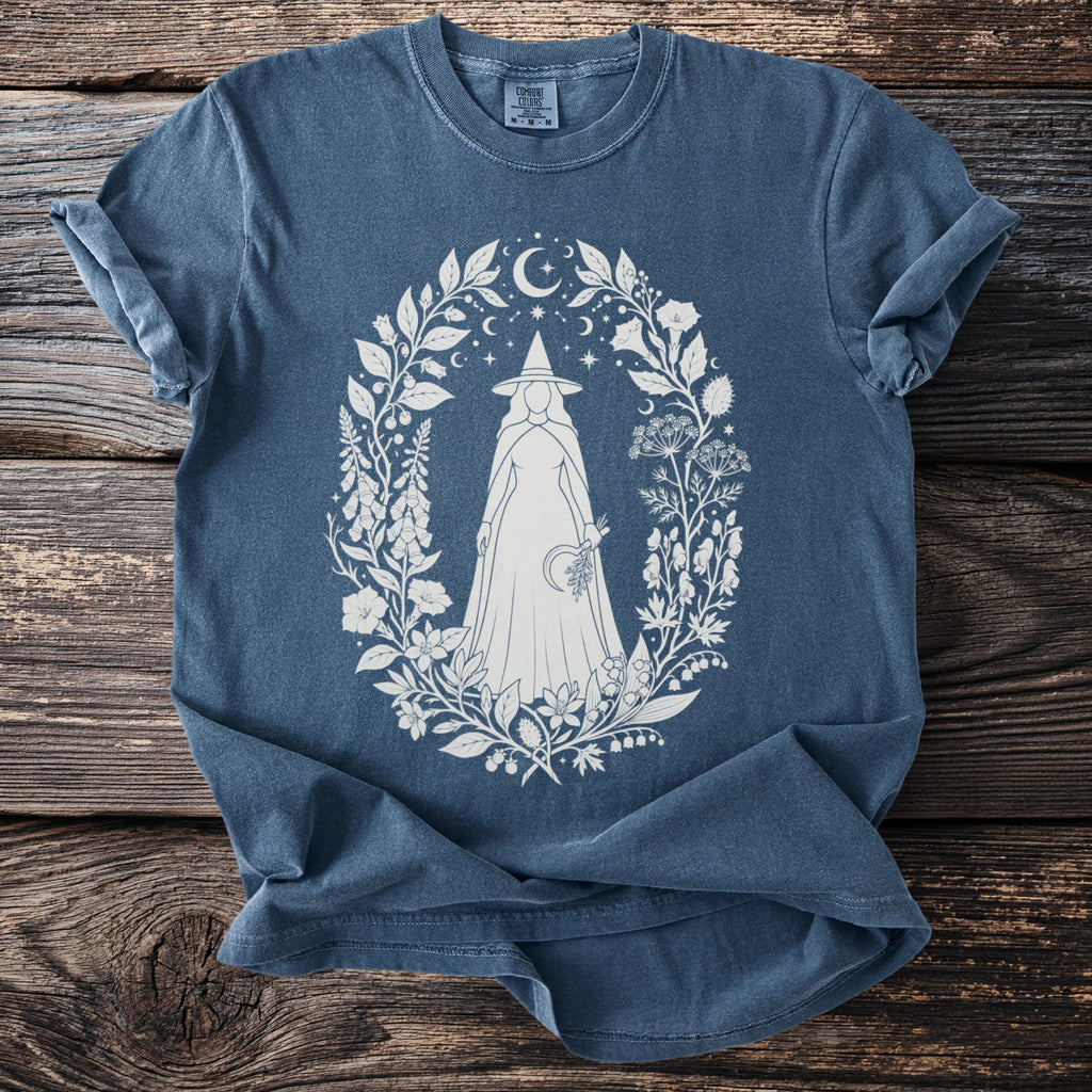 Blue shirt with white witch illustration surrounded by celestial flora and fauna.