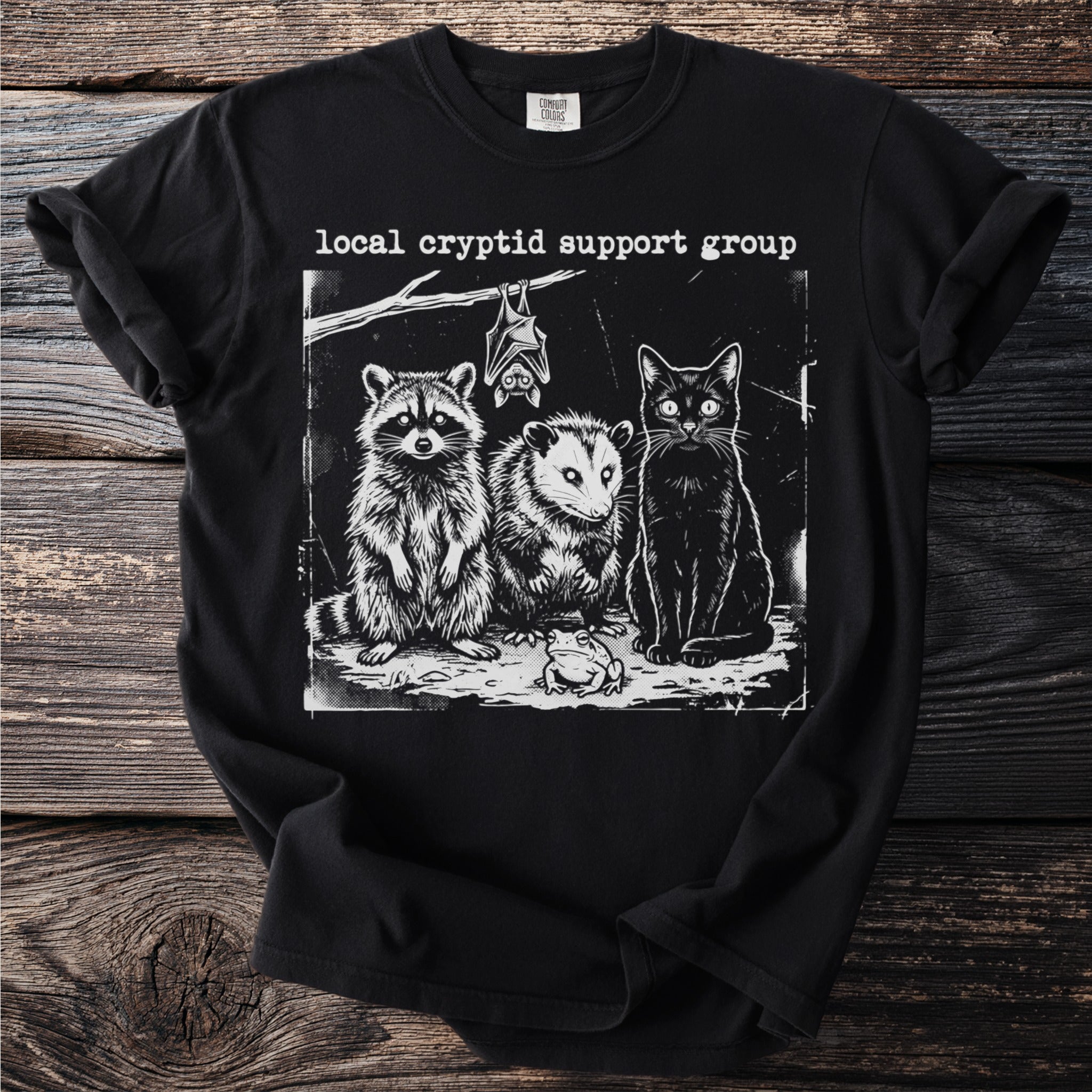 Black t-shirt with "local cryptid support group" text and raccoon, possum, bat, cat, and frog illustration.