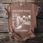Brown t-shirt with "god forbid women have hobbies" and vintage illustration of women in library.