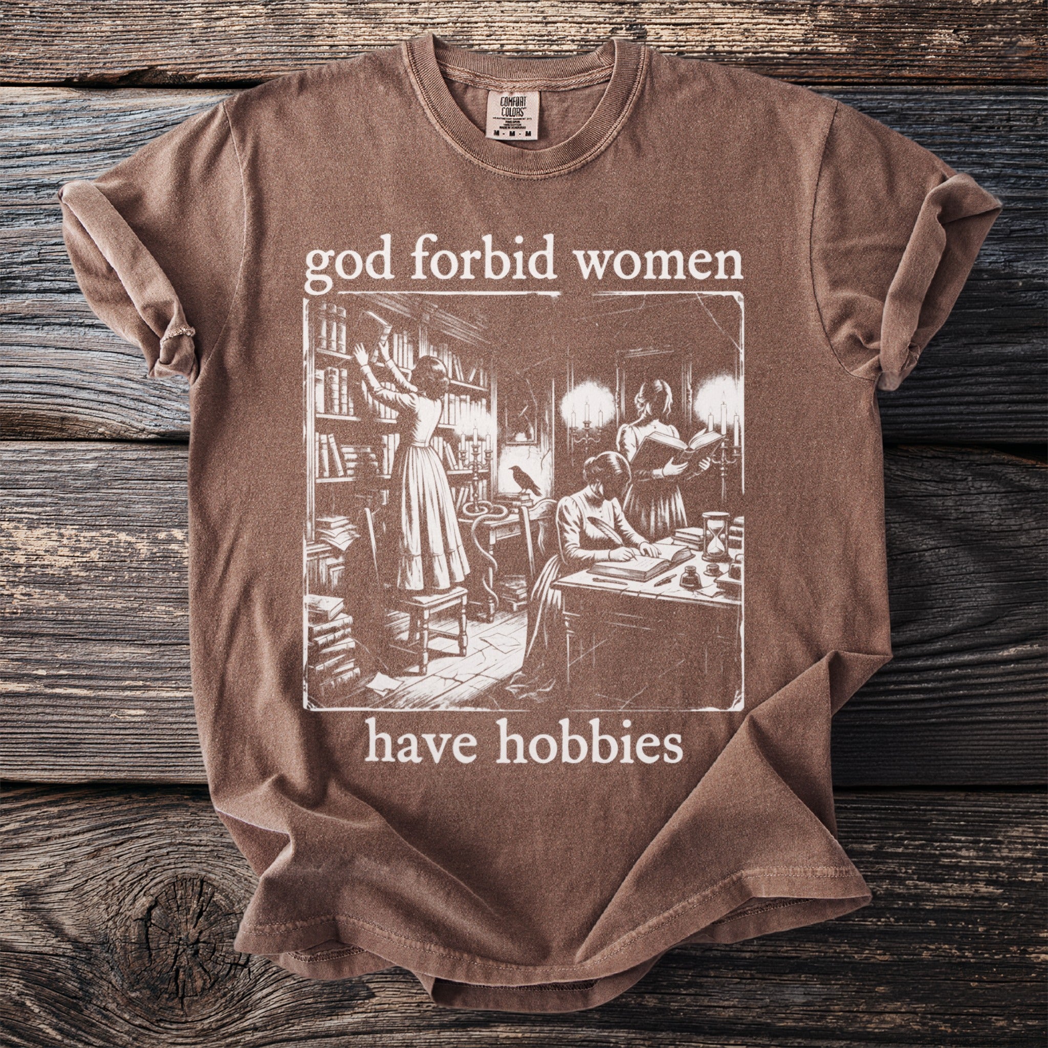 Brown t-shirt with "god forbid women have hobbies" and vintage illustration of women in library.