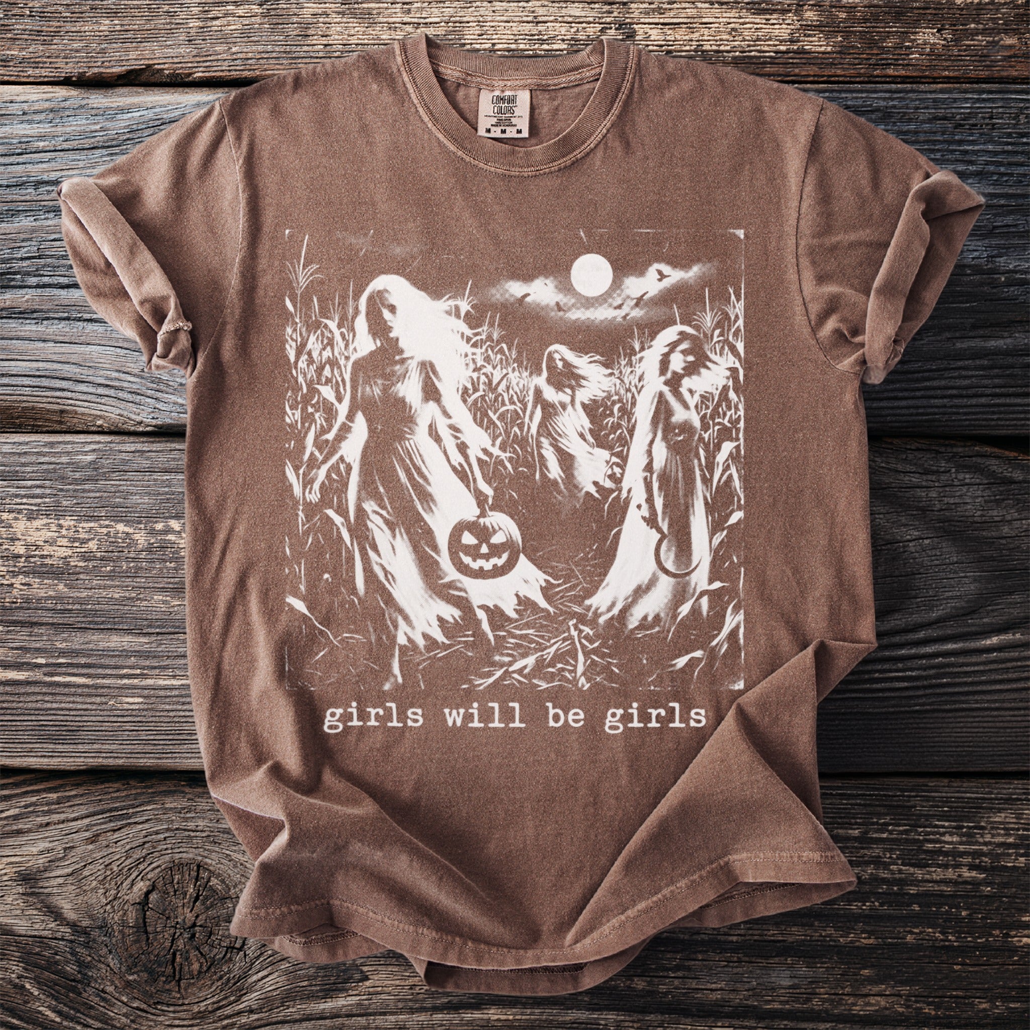 Brown t-shirt with white graphic of figures in cornfield, holding pumpkin and sickle. Text reads "girls will be girls."