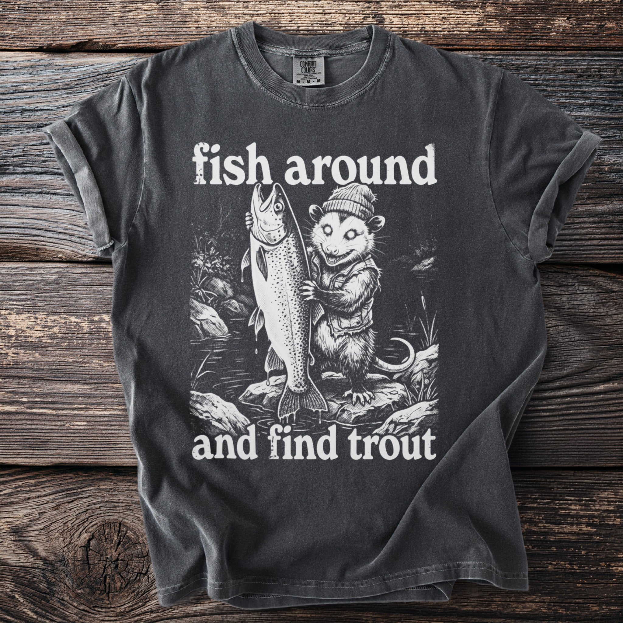 Dark t-shirt graphic with opossum holding large trout and text "fish around and find trout."