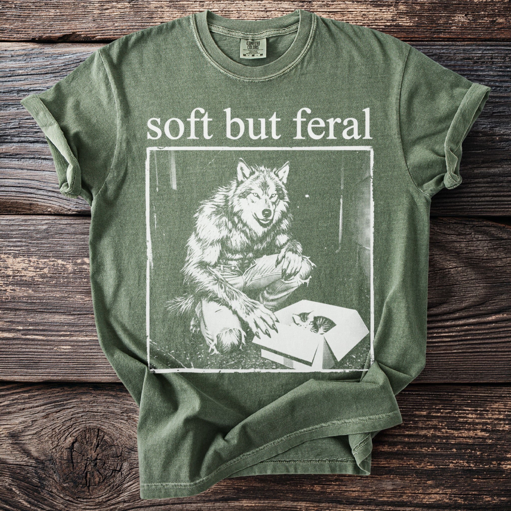 Olive green t-shirt with "soft but feral" text and a werewolf looking into a box with a kitten.