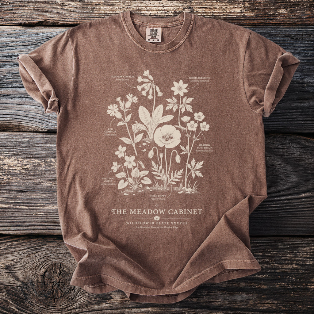Brown t-shirt with a botanical illustration of wildflowers and text "The Meadow Cabinet".