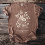 Brown t-shirt with a botanical illustration of wildflowers and text "The Meadow Cabinet".