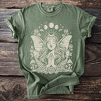 Green t-shirt with a cream graphic of a winged woman with praying hands and celestial elements.