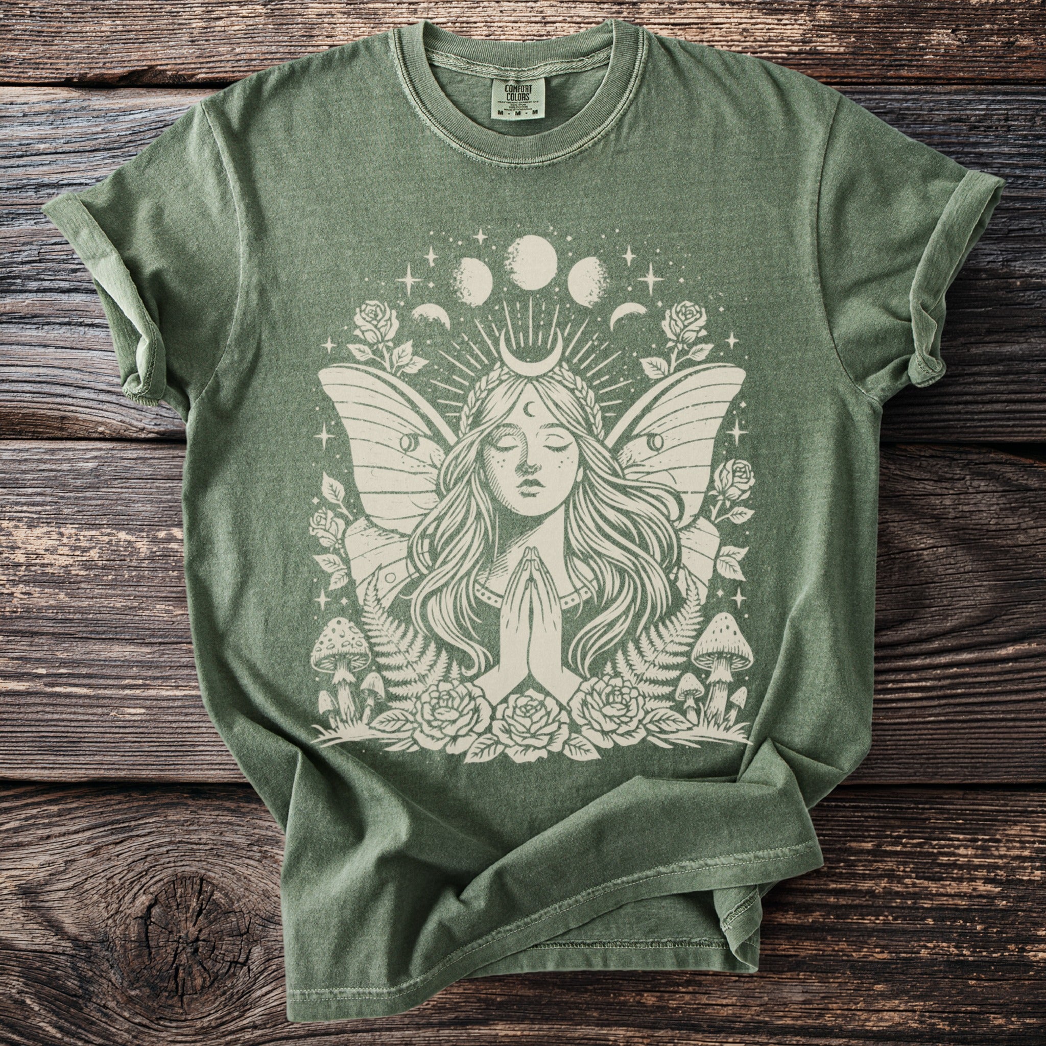 Green t-shirt with a cream graphic of a winged woman with praying hands and celestial elements.
