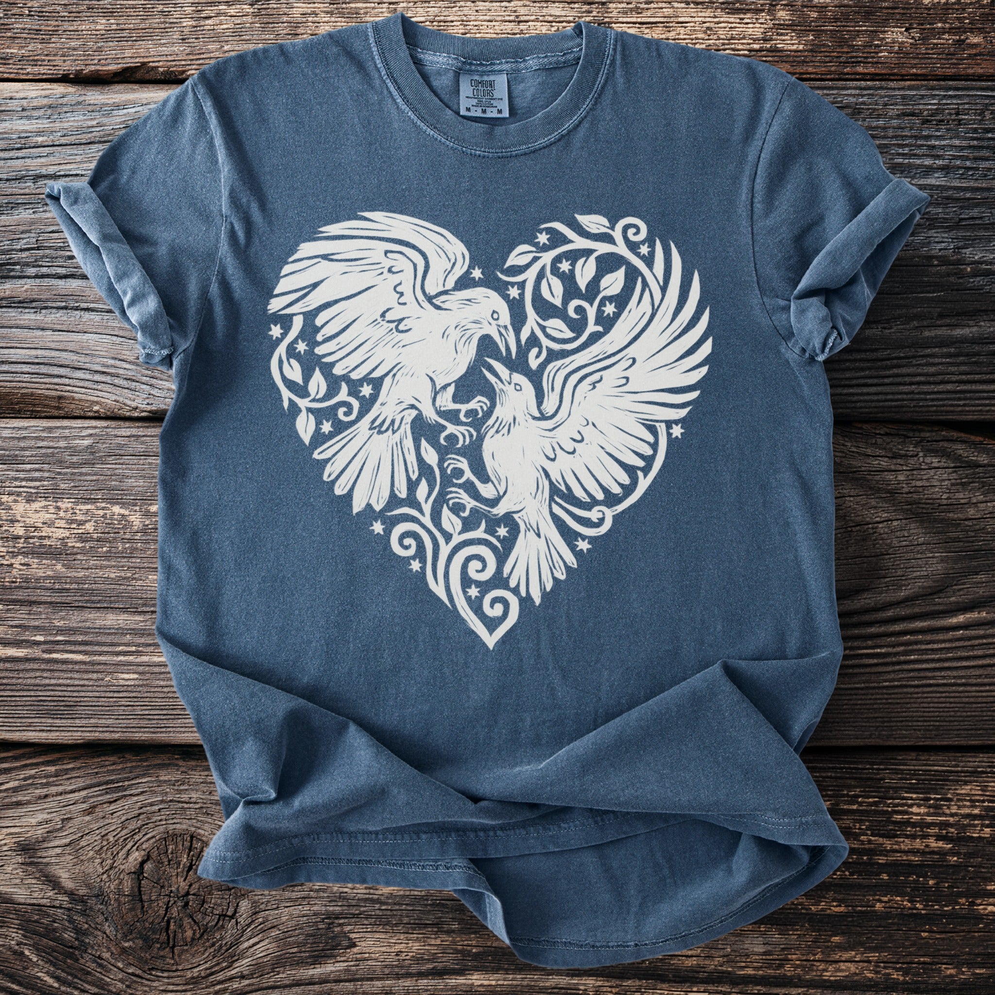 Blue t-shirt with white graphic of two birds in a heart shape on a wooden surface.