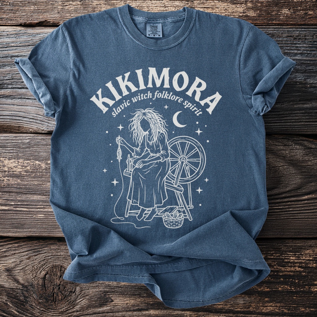 Blue t-shirt with white graphic of a woman spinning yarn and text "KIKIMORA slavic witch folklore spirit".