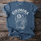 Blue t-shirt with white graphic of a woman spinning yarn and text "KIKIMORA slavic witch folklore spirit".