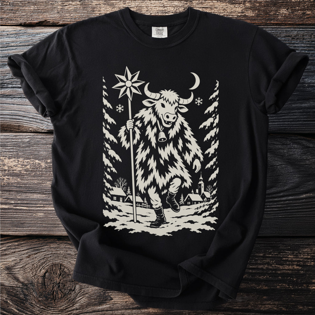 Black t-shirt with a cream folk-cut print: a bull-masked figure in a shaggy coat walks through a snowy village carrying a star on a pole.