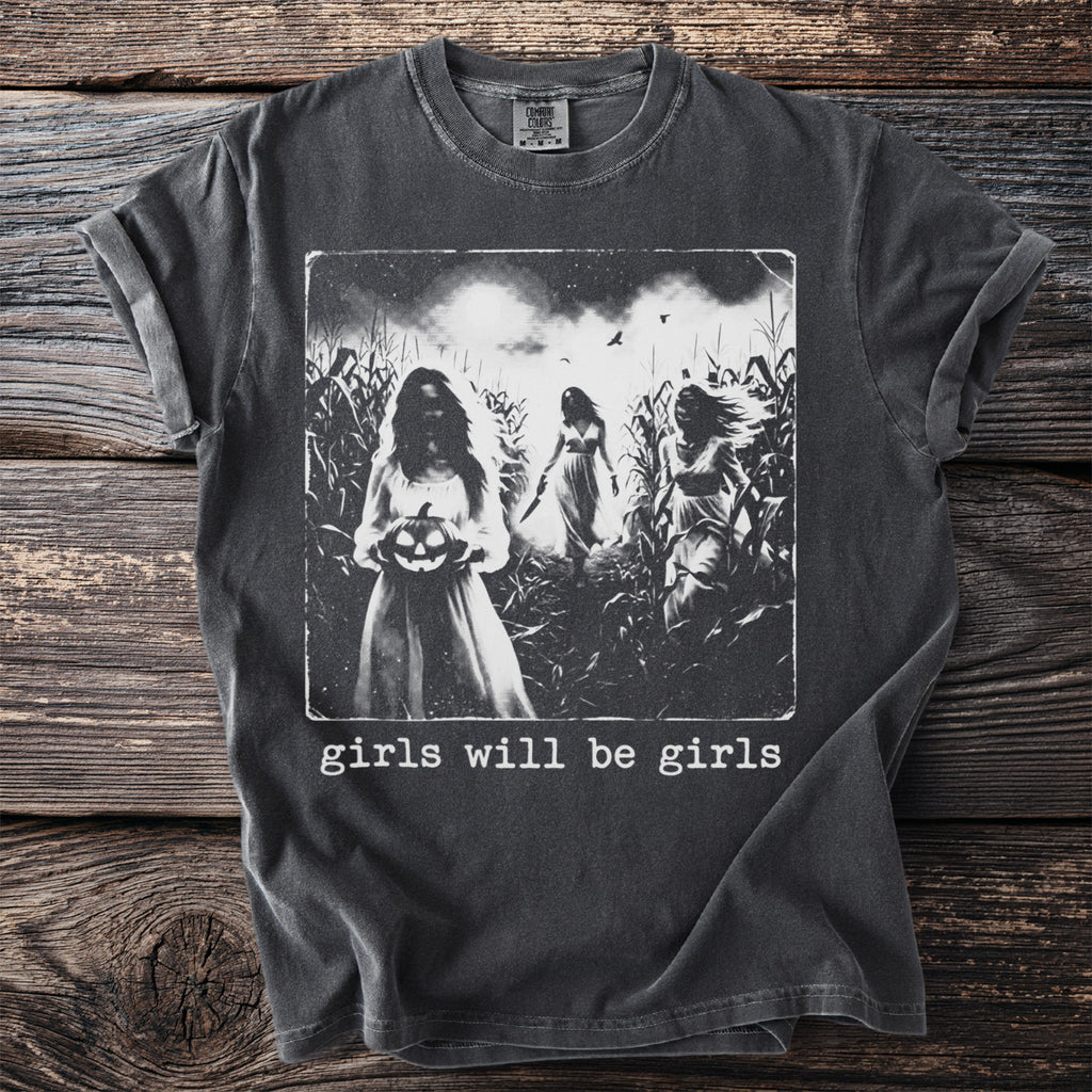 Vintage-style t-shirt graphic of three girls in a cornfield with a pumpkin and knife, text "girls will be girls".