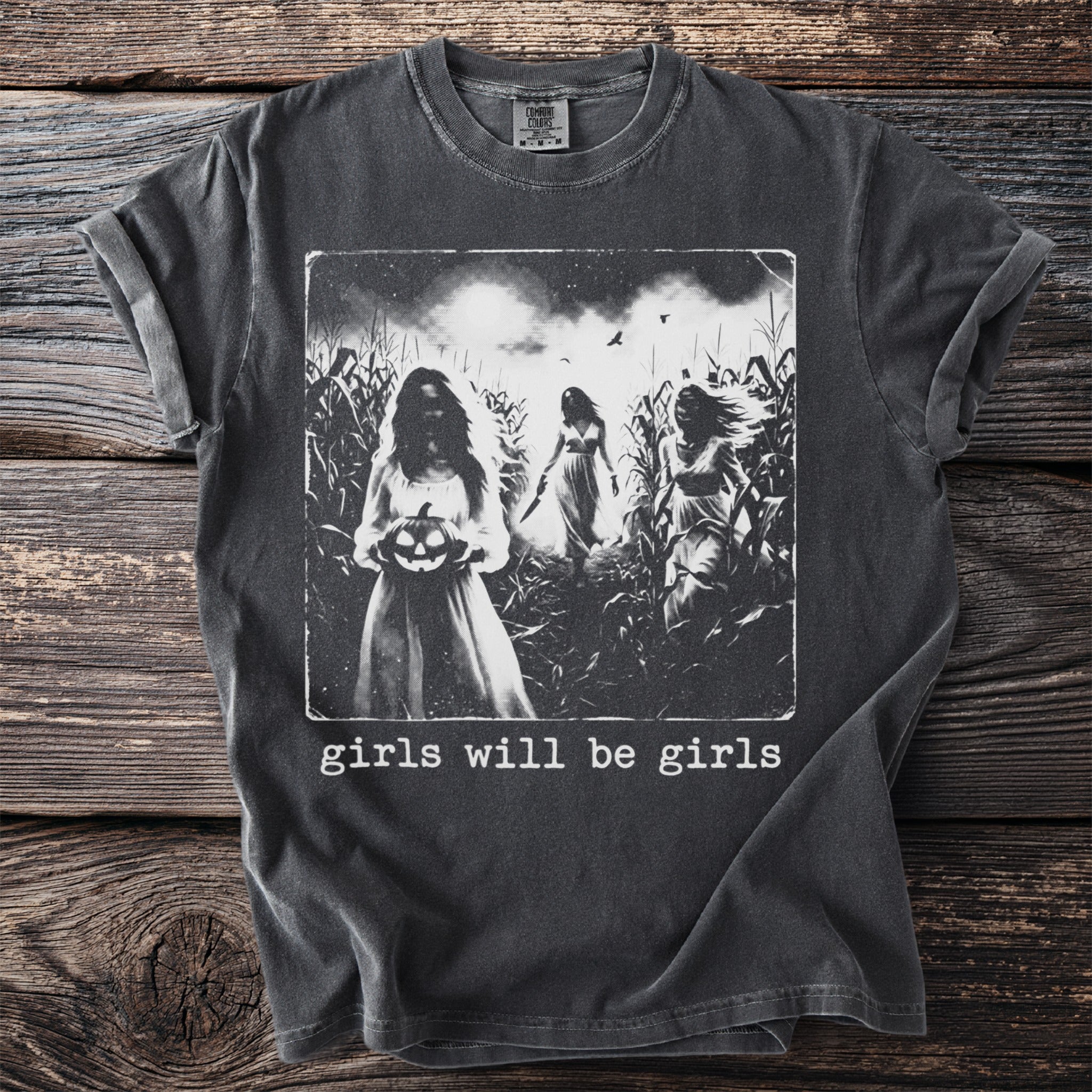Vintage-style t-shirt graphic of three girls in a cornfield with a pumpkin and knife, text "girls will be girls".