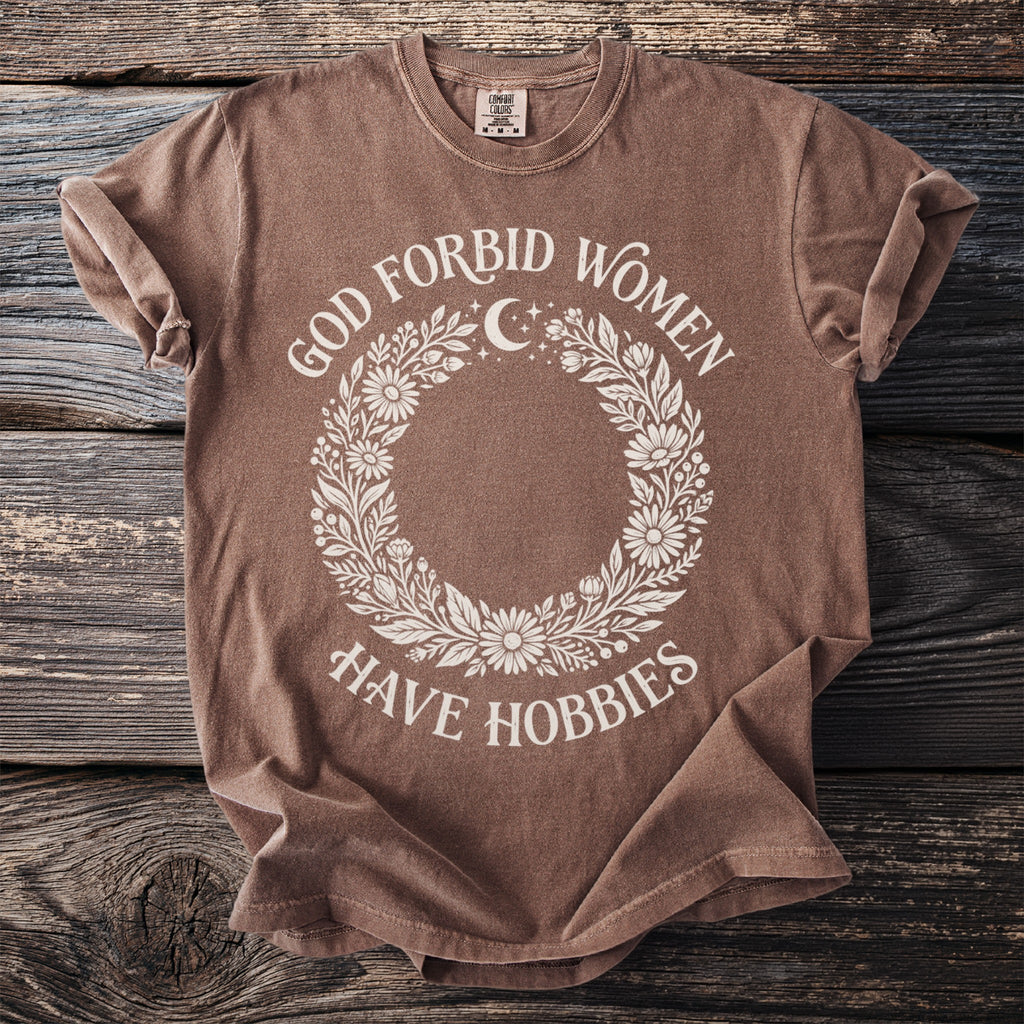 Brown t-shirt with "God forbid women have hobbies" text and floral wreath design.