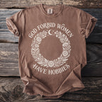 Brown t-shirt with "God forbid women have hobbies" text and floral wreath design.