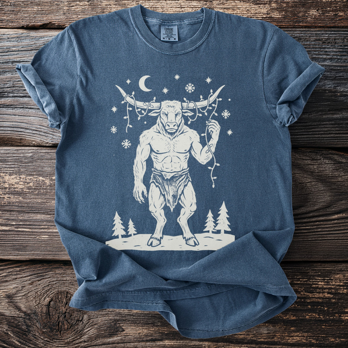 Denim blue t-shirt with a cream folk-cut print: the Minotaur stands in the snow with a string of Christmas lights tangled around his horns.