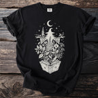 Black t-shirt with a white illustration of a witch emerging from an open book surrounded by plants.