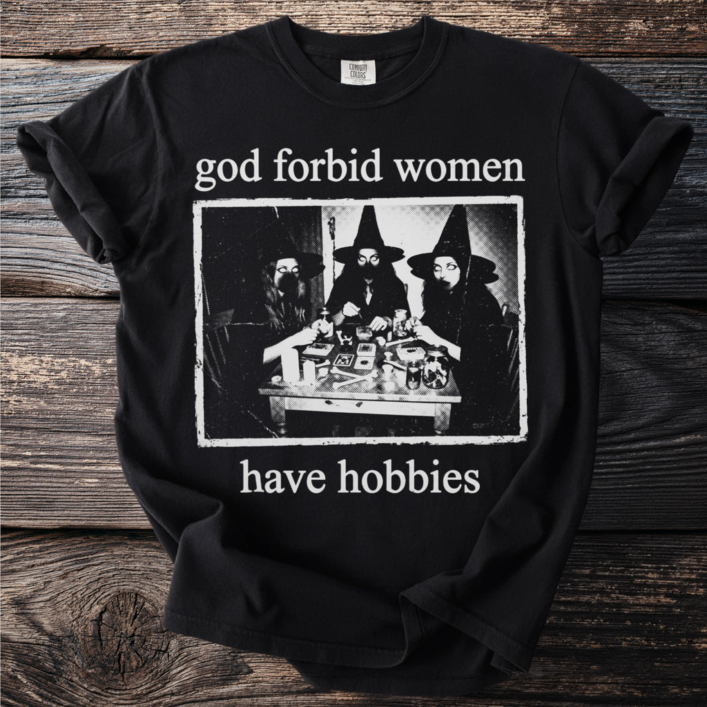 Black t-shirt with text "god forbid women have hobbies" and a B&W image of witches.