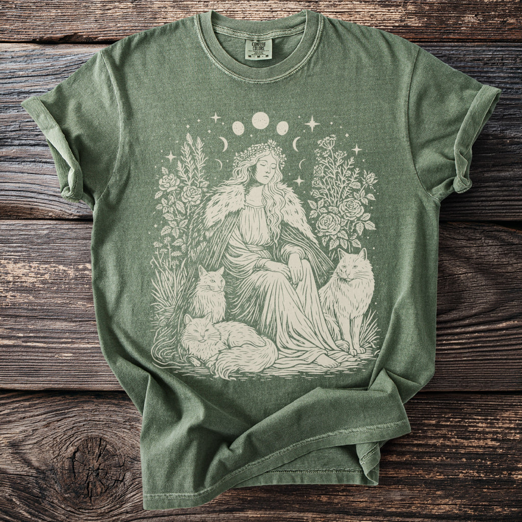 Green t-shirt with a cream design of a woman in a floral crown with two cats and celestial elements.
