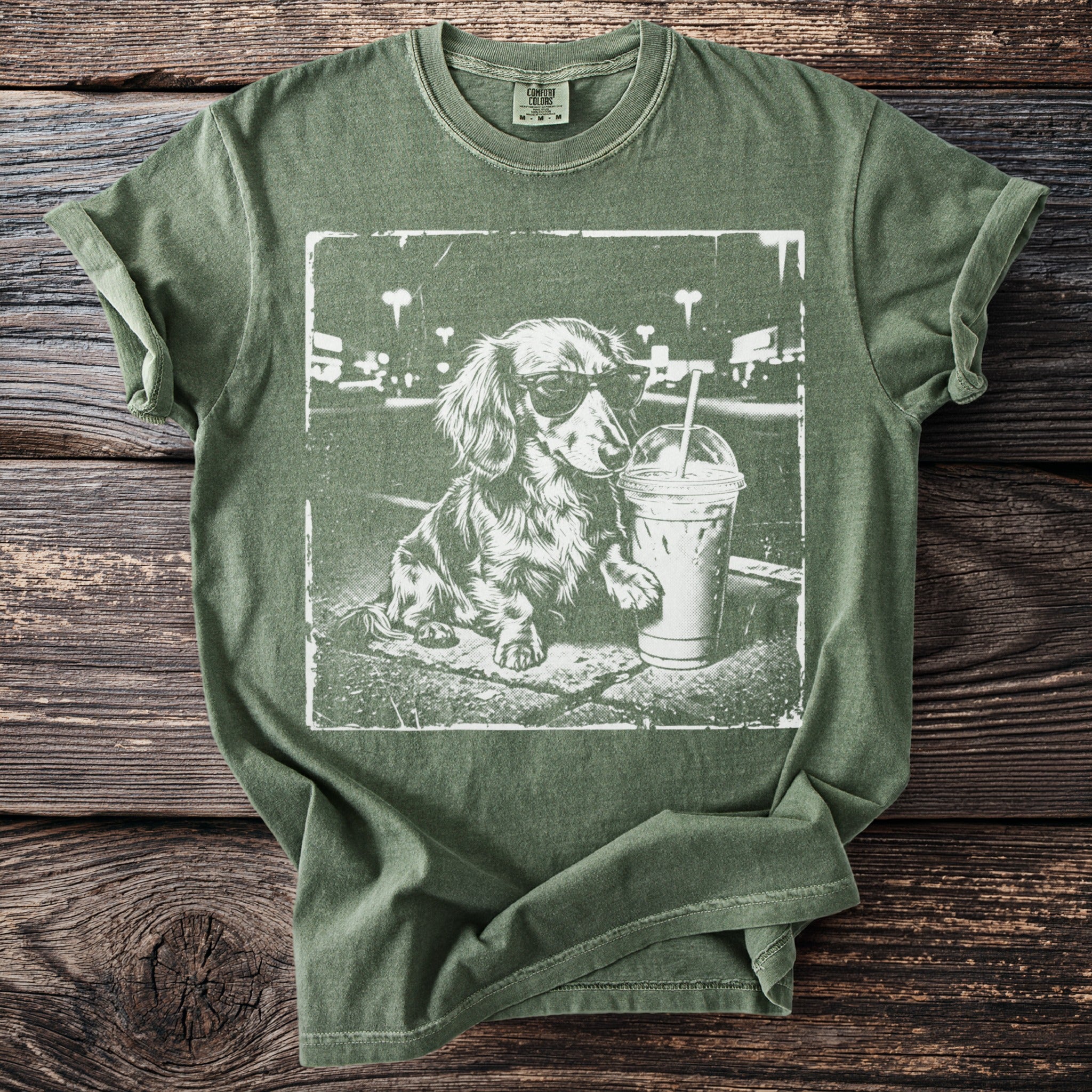 Green t-shirt with a distressed print of a dachshund wearing sunglasses next to a drink.