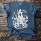 Blue t-shirt with a white graphic of witches, herbs, and potions surrounded by foliage.