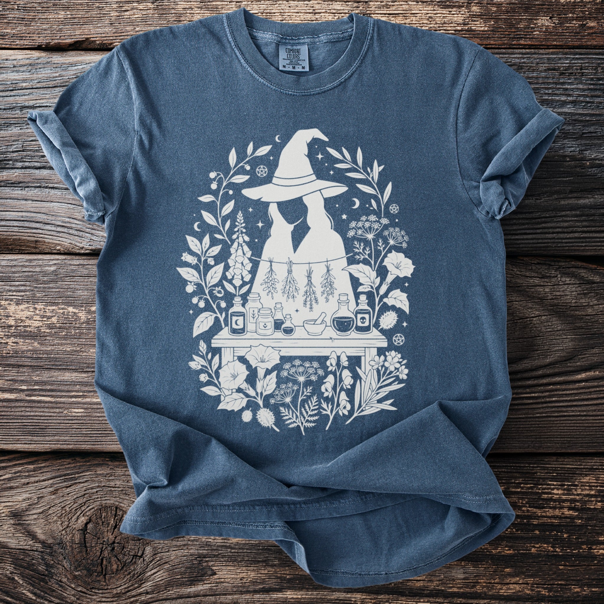 Blue t-shirt with a white graphic of witches, herbs, and potions surrounded by foliage.