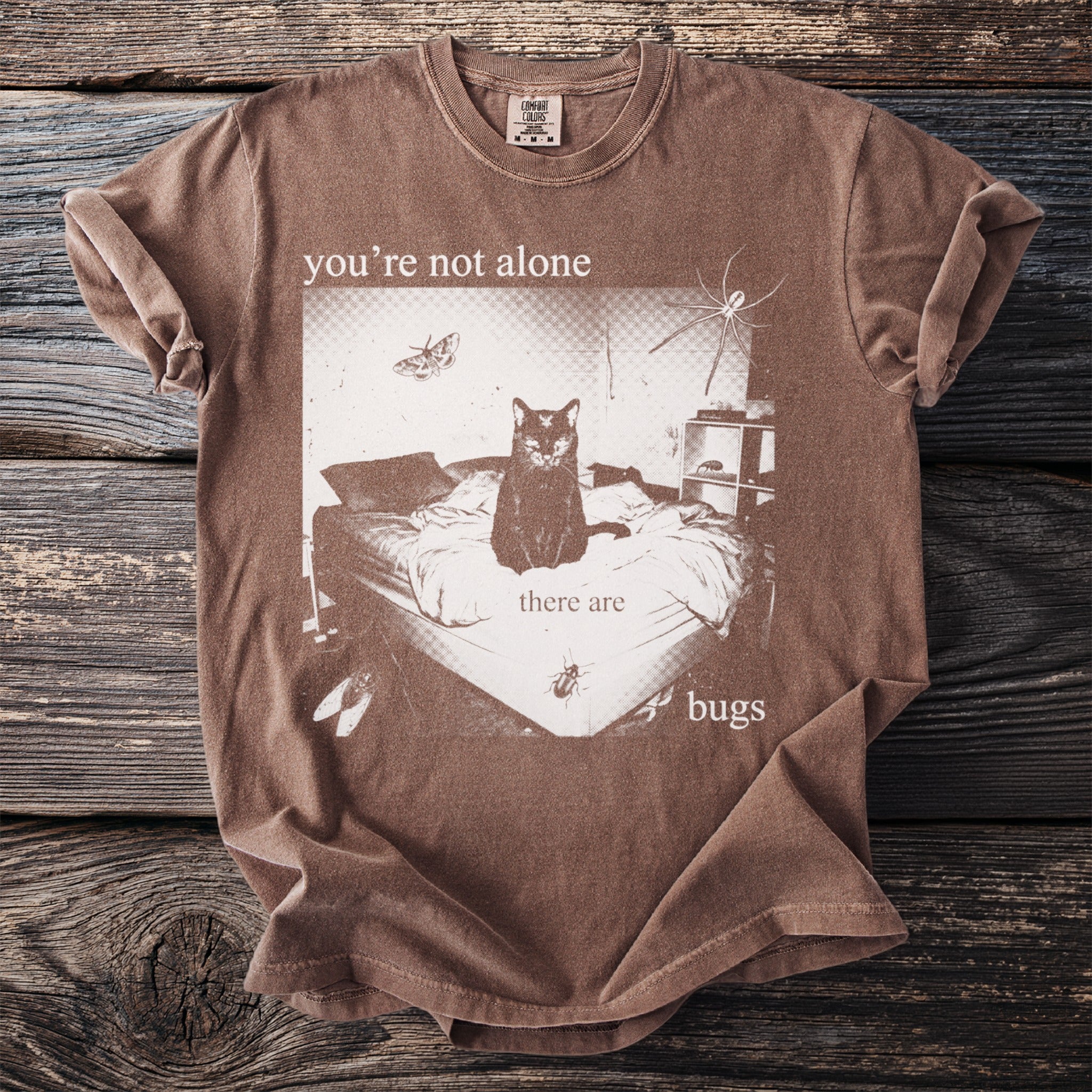 Brown t-shirt with a graphic of a black cat on a bed surrounded by bugs and text.