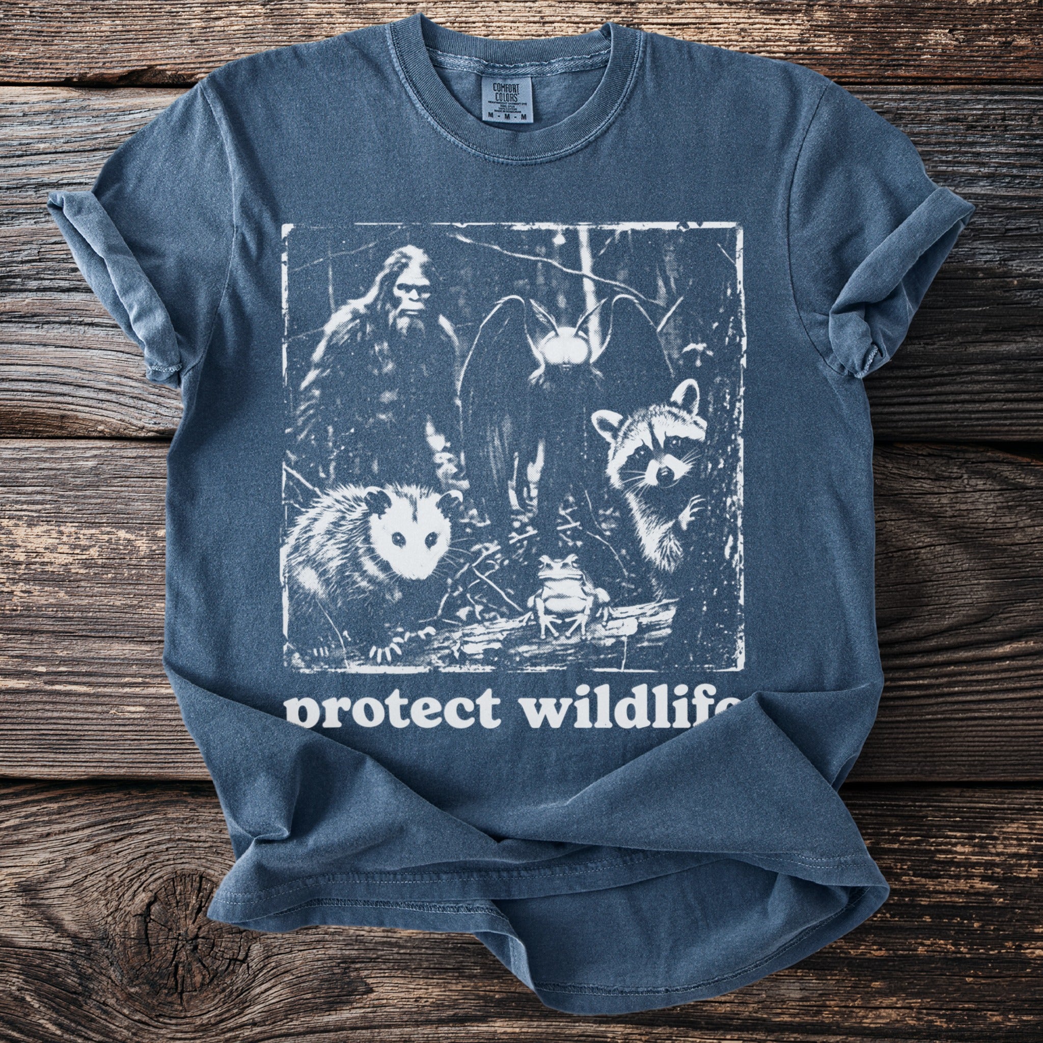 Blue t-shirt graphic features Bigfoot, moth, raccoon, opossum, and frog with "protect wildlife" text.