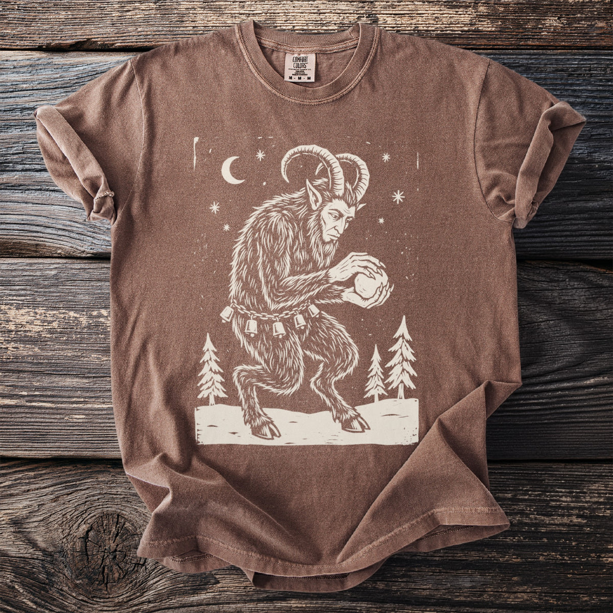 Espresso brown t-shirt with a cream folk-cut print: Krampus crouches among snowy pines, packing a snowball between his clawed hands.