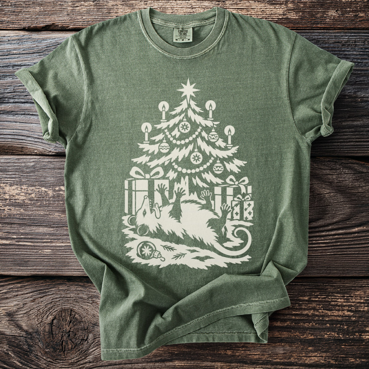 Moss green t-shirt with a cream folk-cut print: an opossum lies on its back under a candlelit Christmas tree, playing dead among the presents.