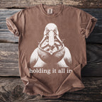 Brown t-shirt with graphic of pelican holding fish, and text "holding it all in."