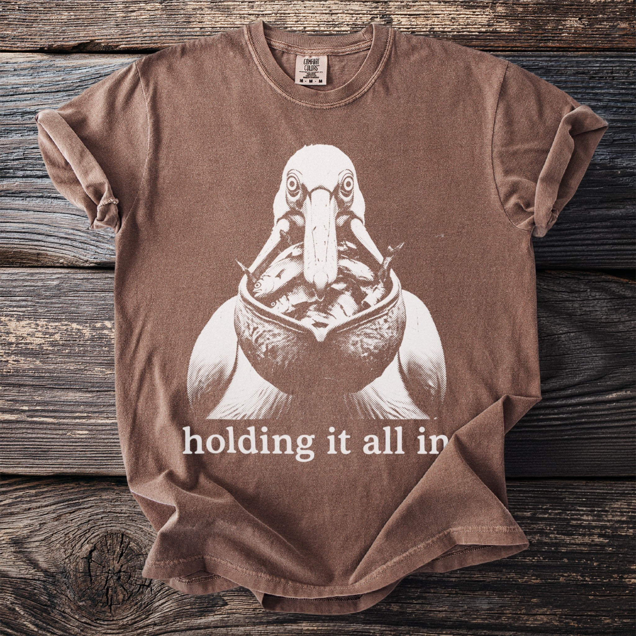 Brown t-shirt with graphic of pelican holding fish, and text "holding it all in."