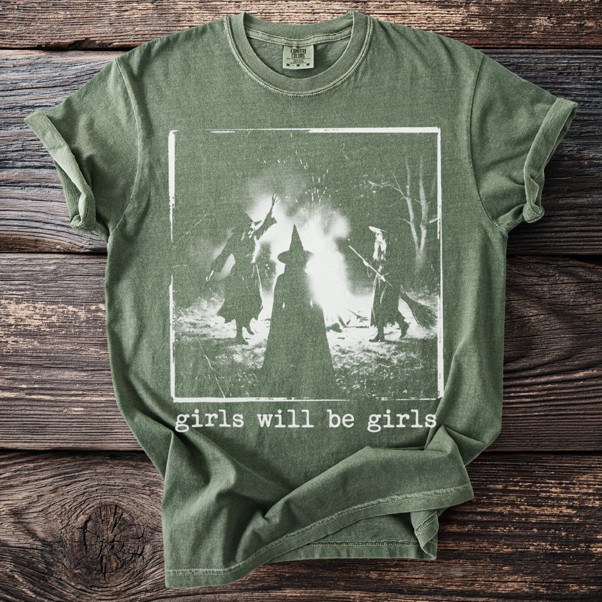 Green t-shirt with three figures in witch hats around a bonfire, text reads "girls will be girls".
