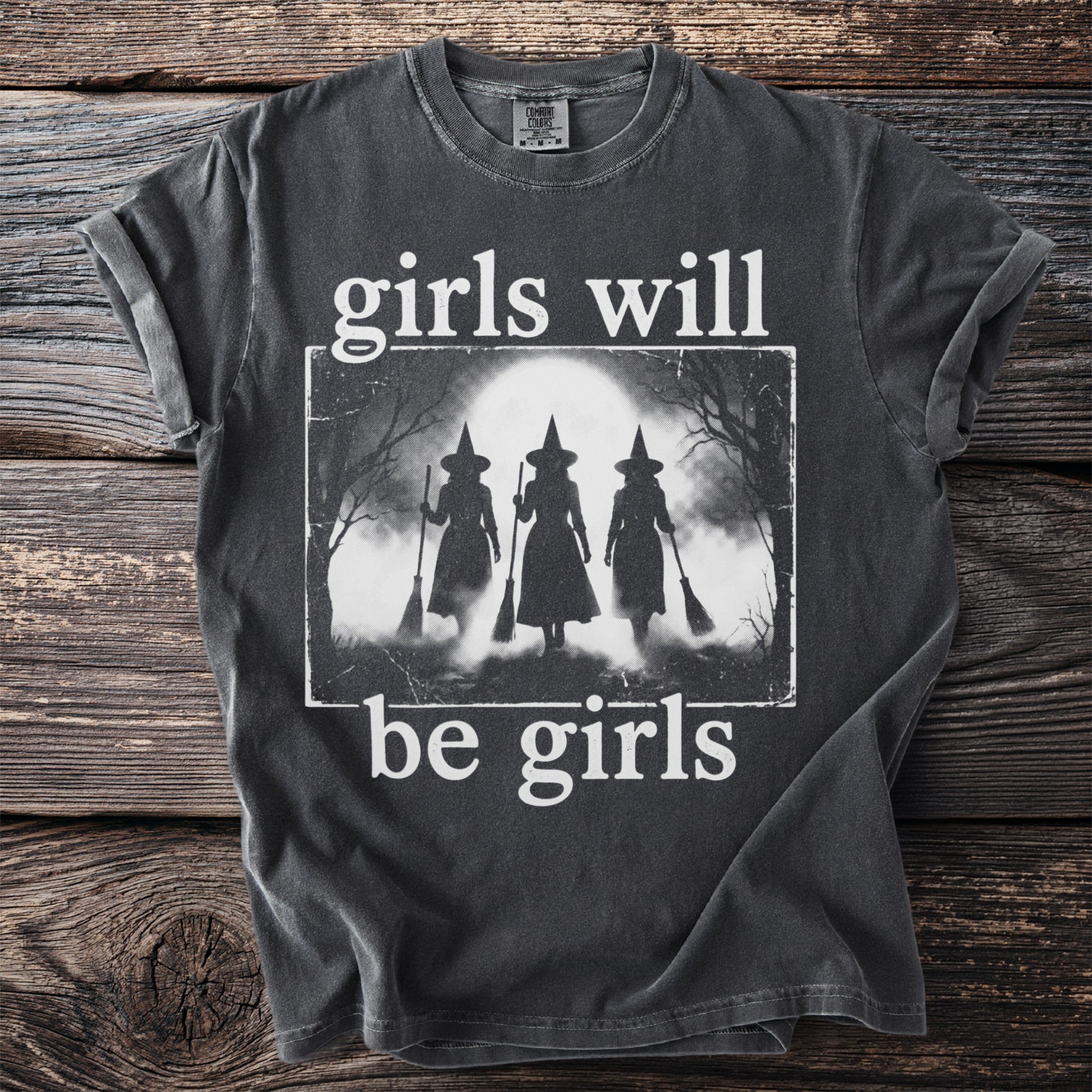 Dark t-shirt with white text "girls will be girls" and silhouette of witches.