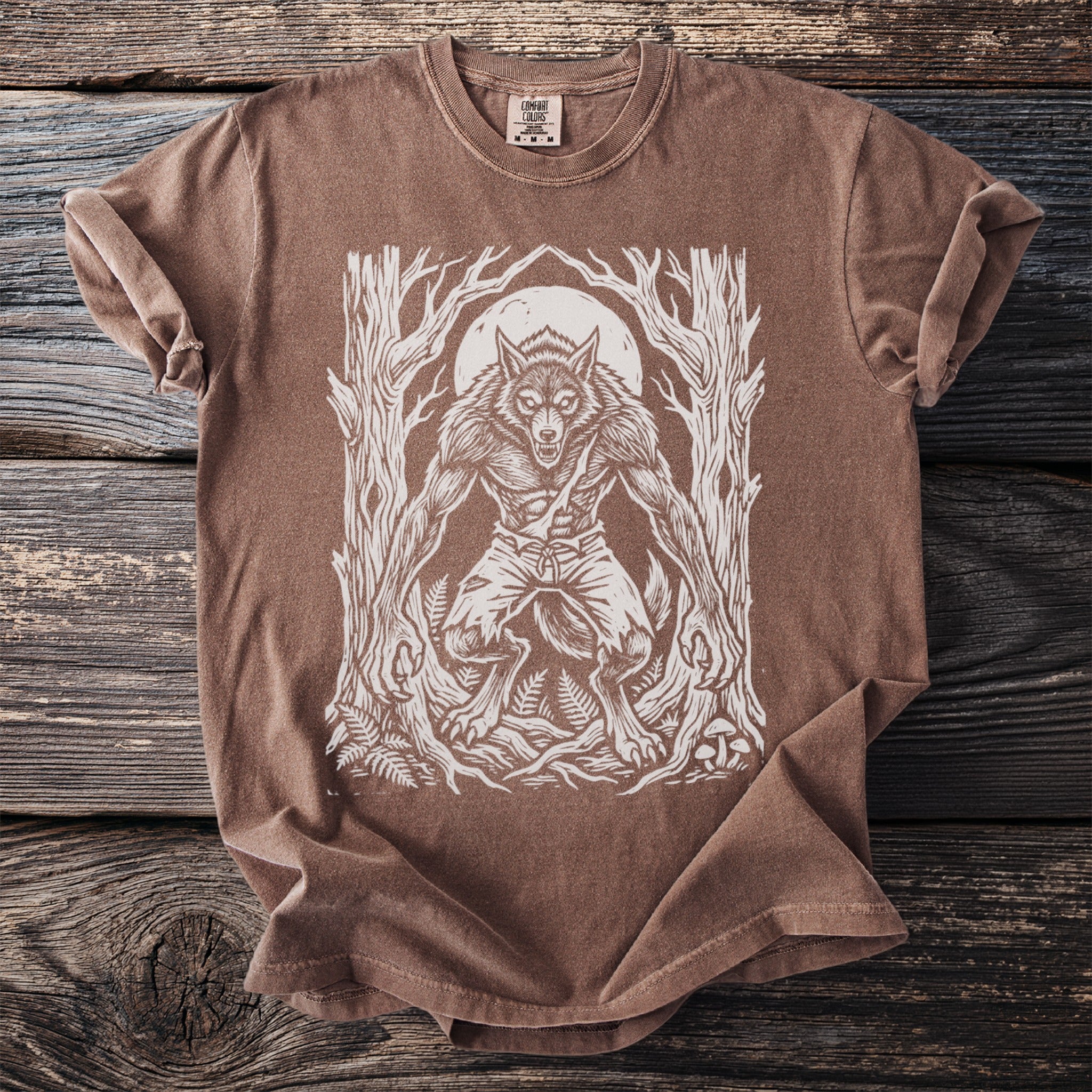 Brown t-shirt with a white linocut-style werewolf and moon graphic in a forest.
