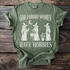 Green t-shirt with white text and illustrations of three witches and a plant.
