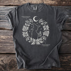 Dark grey t-shirt with white linework of a witch and medicinal plants, moon, and stars.