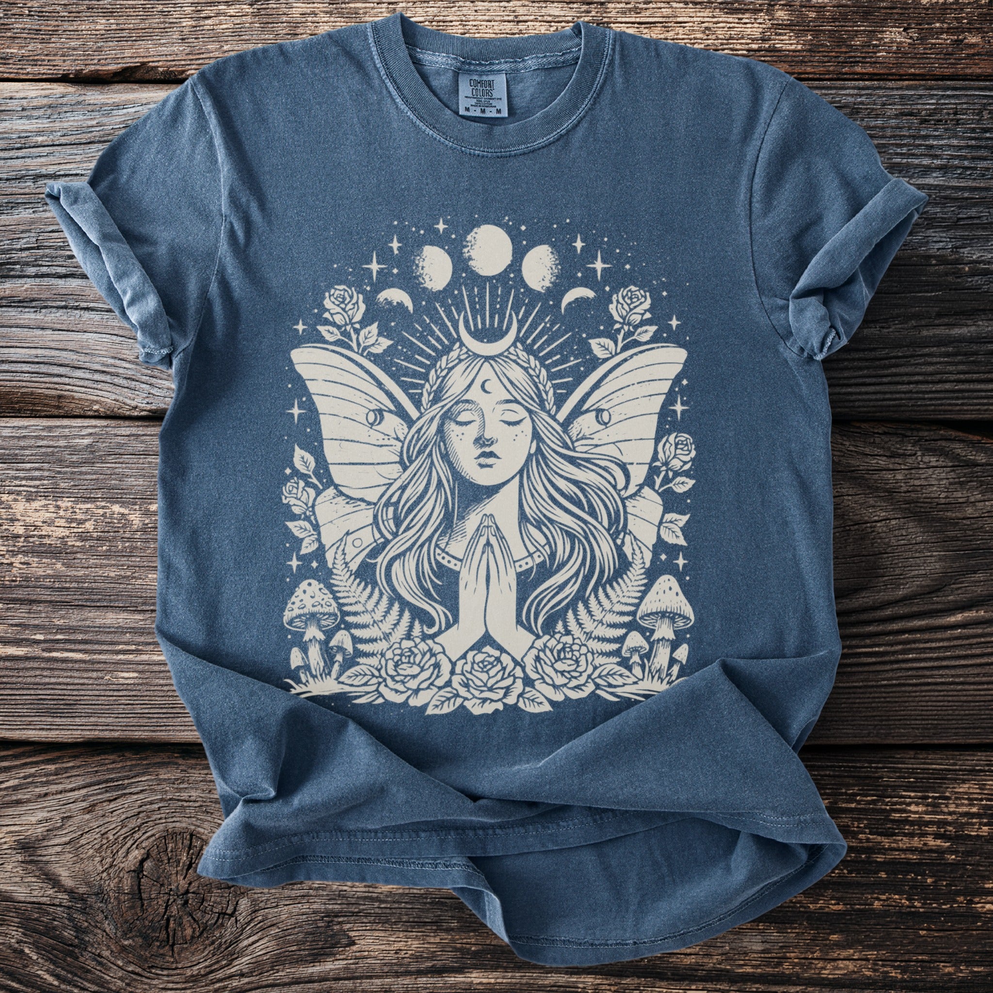 Blue t-shirt with a cream graphic of a woman with moth wings, roses, mushrooms, and moon phases.